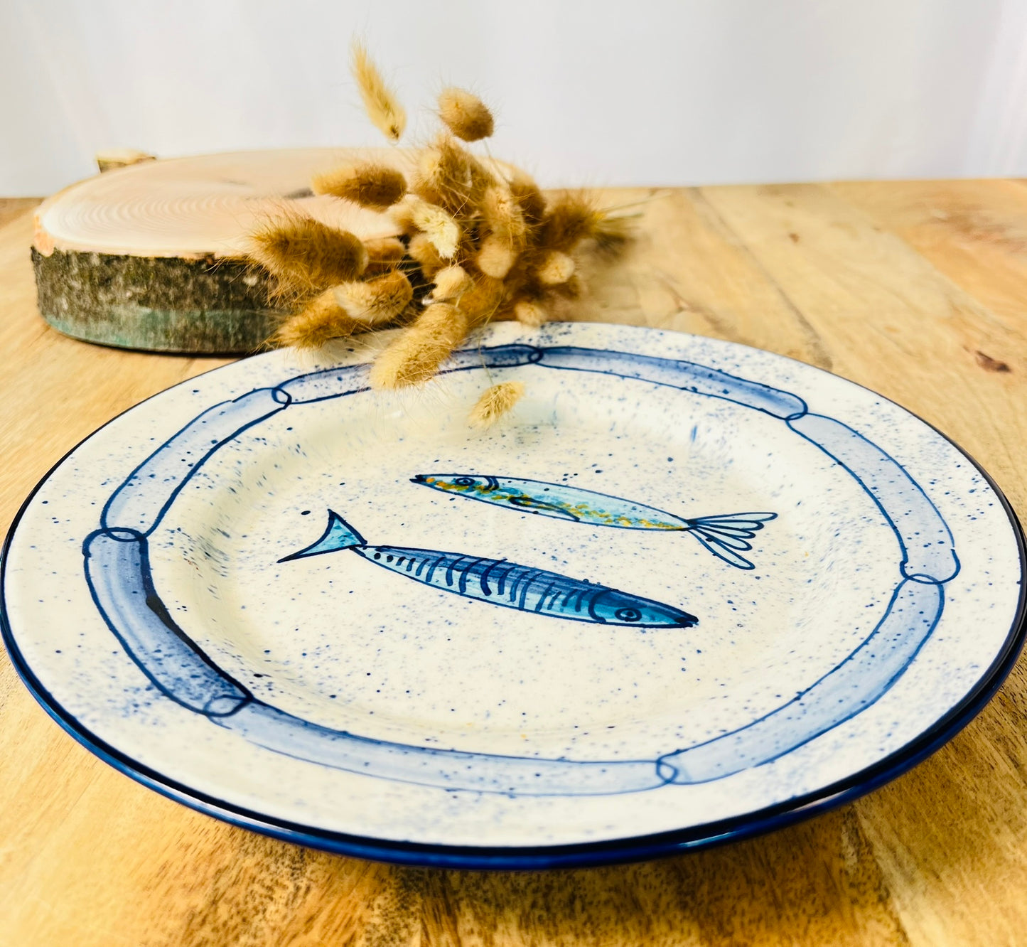 Handgemachter Keramikteller (25 cm) mit handgemaltem Motiv zweier stilisierter Sardinen in verschiedenen Blautönen auf cremeweißem Grund mit blauen Sprenkeln und kettenartigem, blauem Rand.