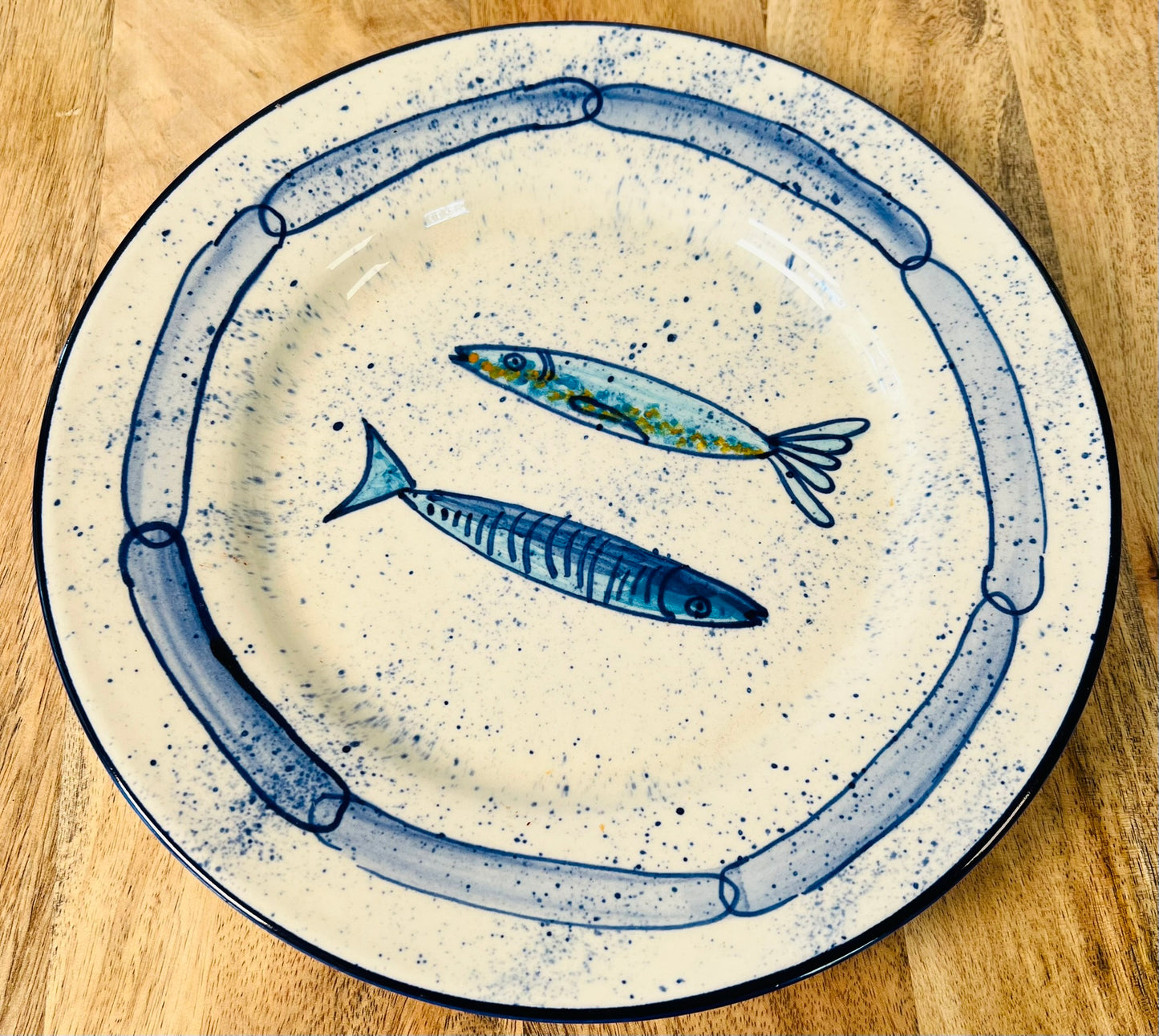 Handgemachter Keramikteller (25 cm) mit handgemaltem Motiv zweier stilisierter Sardinen in verschiedenen Blautönen auf cremeweißem Grund mit blauen Sprenkeln und kettenartigem, blauem Rand.