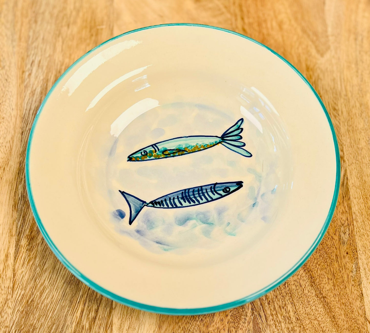 Handgemachter, tiefer Keramikteller (23 cm) mit türkisfarbenem Rand und handgemaltem Motiv zweier stilisierter Fische in Blautönen auf weißem Grund.