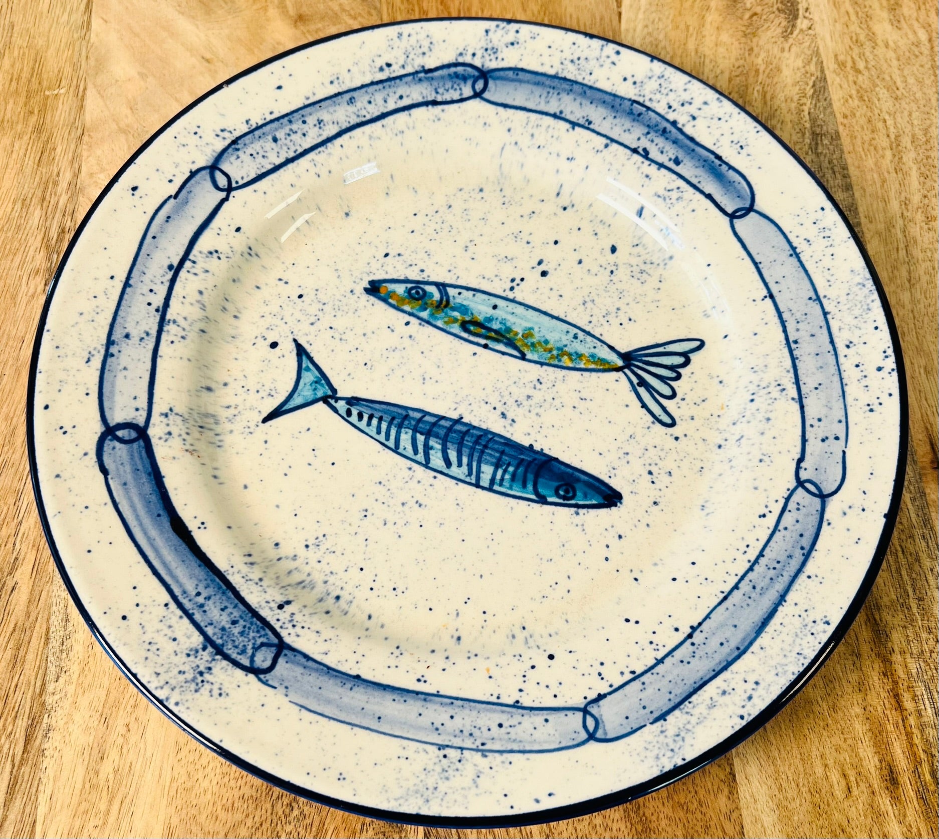 Handgemachter Keramikteller (25 cm) mit handgemaltem Motiv zweier stilisierter Sardinen in verschiedenen Blautönen auf cremeweißem Grund mit blauen Sprenkeln und kettenartigem, blauem Rand.