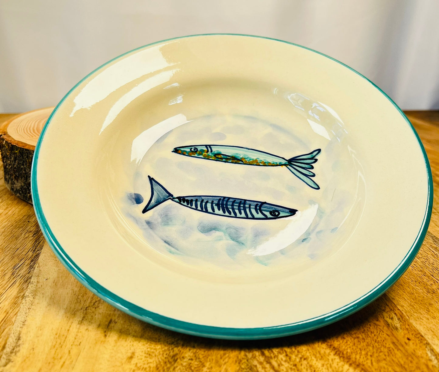 Handgemachter, tiefer Keramikteller (23 cm) mit türkisfarbenem Rand und handgemaltem Motiv zweier stilisierter Fische in Blautönen auf weißem Grund.