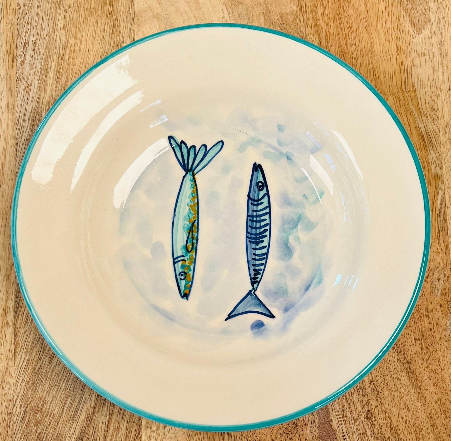 Handgemachter, tiefer Keramikteller (23 cm) mit türkisfarbenem Rand und handgemaltem Motiv zweier stilisierter Fische in Blautönen auf weißem Grund.